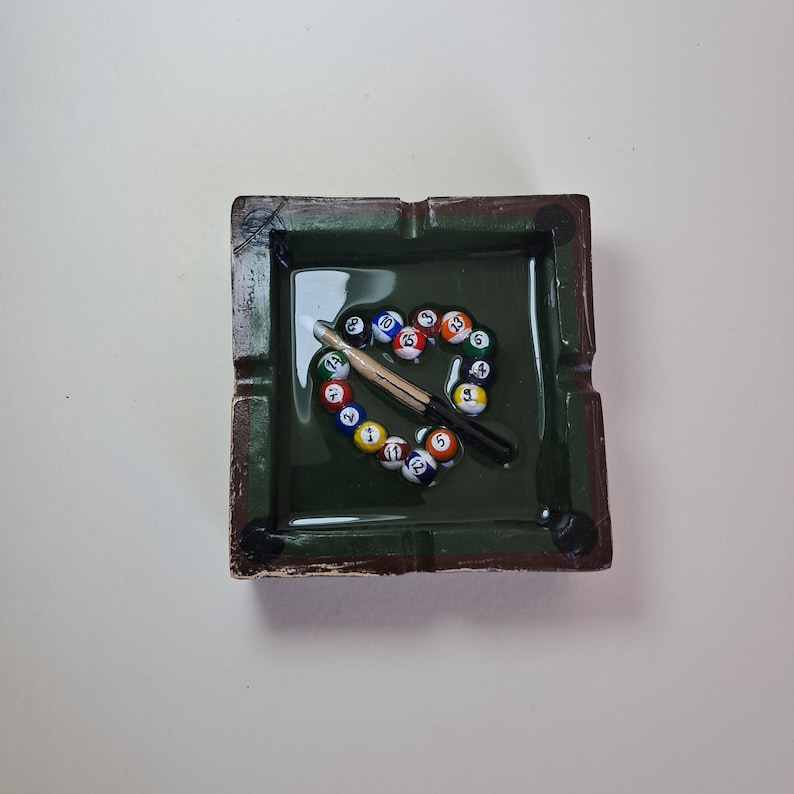 May include: A square, green ashtray with a brown rim. Inside, a heart shape is formed by colorful billiard balls and a cue stick. The ashtray has a glossy finish and is designed to resemble a pool table.