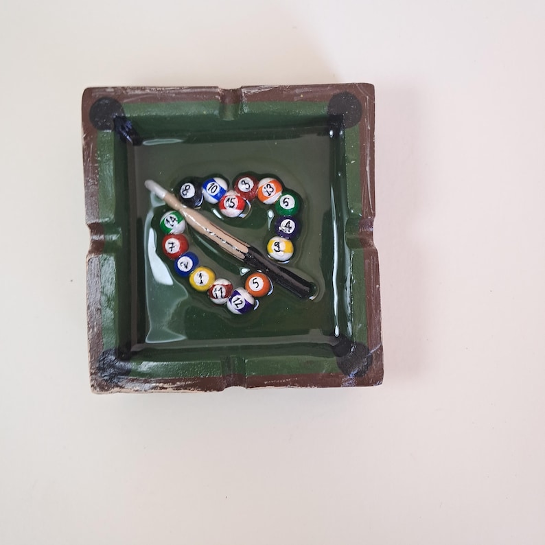 May include: A square, green ashtray with a glossy finish. Inside, a miniature pool table scene is depicted with a cue stick and a heart-shaped arrangement of numbered billiard balls. The ashtray has a dark brown border.