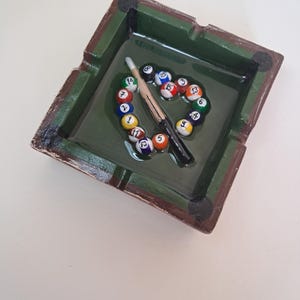 May include: A square ashtray with a green interior and brown trim, featuring a miniature pool table design. The ashtray holds a cue stick and a circle of numbered billiard balls, creating a unique decorative piece.