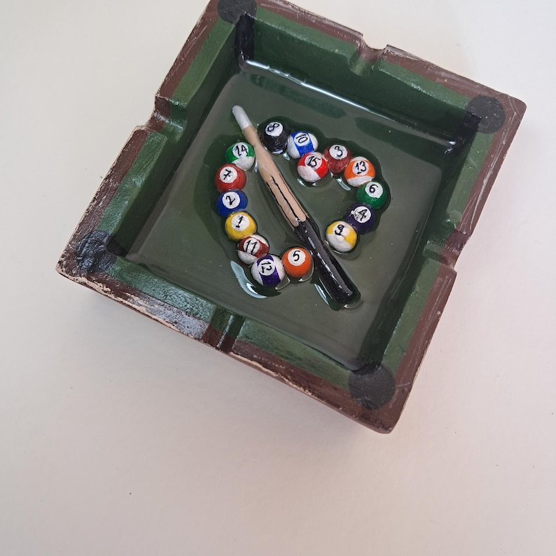 May include: A square ashtray with a green interior and brown trim, featuring a miniature pool table design. The ashtray holds a cue stick and a circle of numbered billiard balls, creating a unique decorative piece.