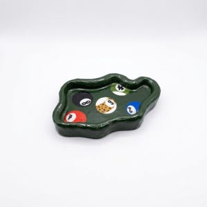 May include: A dark green, irregularly shaped ceramic tray featuring a glossy finish. The interior surface is decorated with five circular pool ball designs, including a red seven ball, a black eight ball, a leopard print nine ball, a blue two ball, and a green ball.