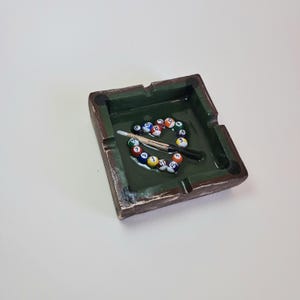 May include: A square ashtray with a green interior and brown exterior, resembling a miniature pool table. The ashtray contains a set of billiard balls, a cue stick, and a cue chalk. The ashtray is a unique decorative item.