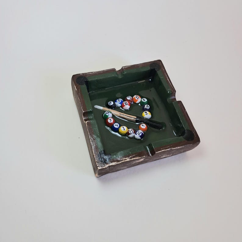 May include: A square ashtray with a green interior and brown exterior, resembling a miniature pool table. The ashtray contains a set of billiard balls, a cue stick, and a cue chalk. The ashtray is a unique decorative item.