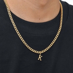 Peut inclure: Un collier à chaîne gourmette doré avec un pendentif en forme de lettre K. Le collier est porté autour du cou sur un t-shirt noir uni. La chaîne en métal est composée de maillons entrelacés formant un motif classique et uniforme sur toute la longueur.