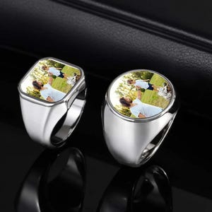 May include: Two silver signet rings, one square and one round, each featuring a color photograph. The photographs depict a person holding a child in the air. The rings are displayed on a reflective black surface.