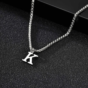 Puede incluir: Collar plateado con un diseño de cadena. El colgante es una letra mayúscula "K". El collar se muestra sobre una superficie negra. La cadena tiene un diseño de eslabones cuadrados pequeños.