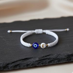 May include: White macrame bracelet with a blue evil eye bead and a white bead with the letter "E". The bracelet has gold-coloured accents and is shown on a dark grey surface.