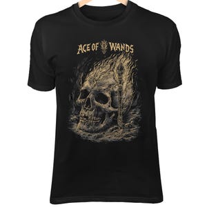 May include: Black t-shirt featuring a detailed graphic of a skull engulfed in flames, with a staff and the text "ACE OF WANDS" in a stylized font. The design is in shades of beige and tan.