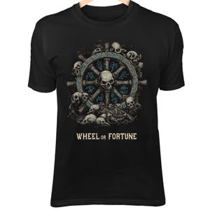 Può includere: T-shirt nera con una grafica dettagliata di un timone decorato con teschi, circondato da teschi e ossa. Il testo "WHEEL OR FORTUNE" è stampato sotto. Il design utilizza tonalità di grigio, verde acqua e bianco.