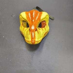 May include: A yellow and orange dinosaur mask with mesh eye coverings and a strap. The mask features a textured surface and is designed to resemble a raptor.