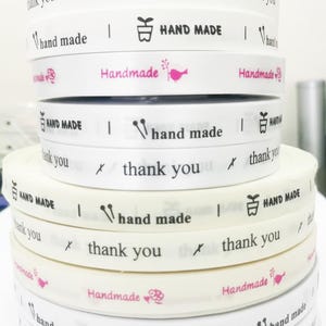 以下が含まれることがあります： 様々なデザインが印刷された白いリボンのロールのスタックです。「Handmade」、「thank you」、「hand made」の文字が含まれています。ピンクの鳥や花のグラフィックが描かれたリボンもあります。クラフトやギフト包装に適しています。