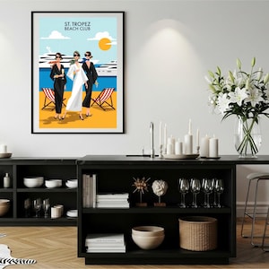 Puede incluir: Una impresión artística enmarcada con el texto "ST. TROPEZ BEACH CLUB", que representa a tres figuras en una playa. La ilustración muestra una escena de playa soleada con un yate, tumbonas y un sol amarillo brillante.