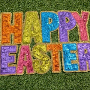 Könnte beinhalten: Ein farbenfrohes, mit Pailletten besetztes "HAPPY EASTER" Schild mit goldenem Rand, vor einem grünen, strukturierten Hintergrund. Die Buchstaben sind in verschiedenen Farben gehalten, darunter Orange, Gelb, Blau, Lila und Rosa.