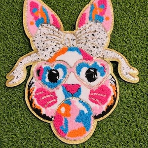 Könnte beinhalten: Ein farbenfroher, skurriler Aufnäher in Form eines Hasengesichts. Der Hase hat herzförmige Brille, eine paillettenbesetzte Schleife und einen goldenen Rand. Das Design weist rosa, blaue, orange und weiße Farben auf.
