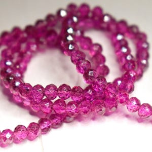 May include: Close-up of a strand of faceted, magenta-colored beads. The beads are strung together, forming a coiled pattern. The beads have a sparkling, crystalline appearance, reflecting light.