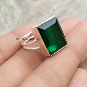 May include: A silver ring with a large, rectangular, emerald-green gemstone. The ring has a double-band design and a polished finish. The text "ShriBalajiJewlry" is visible on the ring.