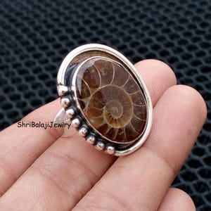 May include: Oval-shaped silver ring featuring a polished, brown fossilized ammonite. The ring has a decorative border of small silver beads and the text "ShriBalajiJewlry".