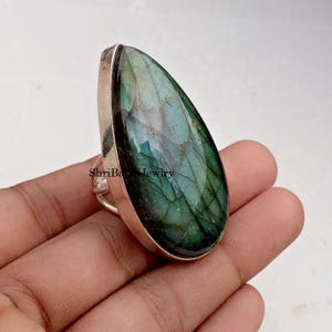 May include: A large, teardrop-shaped ring featuring a polished, iridescent labradorite stone in shades of green, blue, and gray. The stone is set in a silver-colored metal band. The text "ShriBa Jewlry" is visible on the ring.