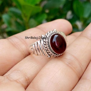 Peut inclure: Une bague en argent avec une pierre précieuse grenat rouge foncé de forme ovale, sertie dans un chaton à texture torsadée. L'anneau présente un motif métallique nervuré avec de petites perles. Le texte ShriBalajiJewelry est visible.