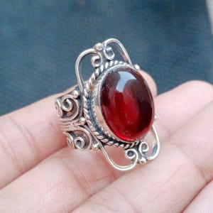 May include: A silver ring featuring a large, oval-shaped, deep red gemstone set in a bezel. The band and setting are decorated with intricate, swirling metalwork and a twisted rope-style border surrounding the central stone.