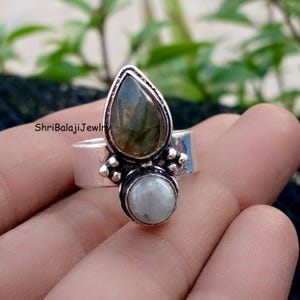 Könnte beinhalten: Ein silberner Ring mit einem birnenförmigen, grün-grauen Edelstein und einem runden, hellfarbigen Stein. Der Ring hat ein breites Band und dekorative Akzente. Der Text "ShriBalajiJewlr" ist sichtbar.