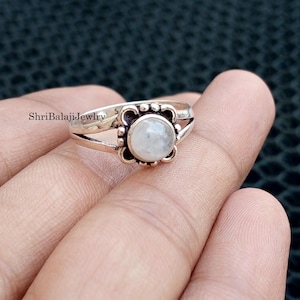 Könnte beinhalten: Ein silberner Ring mit einem runden, hellfarbenen Stein in einer dekorativen Lünette. Der Ring hat ein geteiltes Schaftdesign und der Text "ShriBalajiJewlry" ist auf dem Band sichtbar.