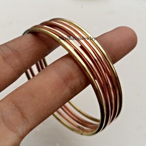 May include: A set of gold, copper, and bronze bangles. The bangles are stacked together, with a mix of gold, copper, and bronze tones. The bangles are held by a person's fingers. The text "ShriBalajiJewlry" is visible on the bangles.