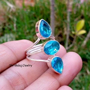 May include: A silver ring featuring three bright blue faceted gemstones in a tiered arrangement. The top stone is pear-shaped, the middle is oval, and the bottom is pear-shaped. The band is made of polished silver wire. Text reads ShriBalajiJewlry.