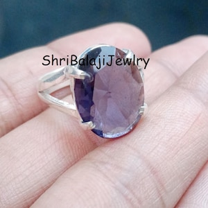 Op de afbeelding: Een zilveren ring met een grote, ovaal geslepen, doorschijnende paarse edelsteen die door vier pootjes wordt vastgehouden. De ringband heeft een gespleten ontwerp. De tekst ShriBalajiJewelry is in zwarte letters zichtbaar bovenin de afbeelding.