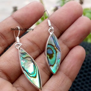 Peut inclure: Boucles d'oreilles en argent avec des coquillages d'ormeau irisés en forme de goutte d'eau. La coquille présente des nuances de vert, de bleu et de violet. Les boucles d'oreilles ont un design simple en forme de crochet. Le texte "ShriBalajiJewlry" est visible.