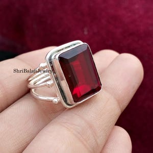 Puede incluir: Un anillo de plata con una gran gema rectangular de color rojo oscuro. El anillo tiene un diseño de doble banda con pequeñas esferas decorativas. El texto "ShriBalajiJewlry" es visible en el anillo.