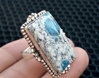 May include: A rectangular silver ring featuring a large, light gray stone with blue and white patterns. The stone is set in a silver frame with a beaded border. The ring is a statement piece of jewelry.