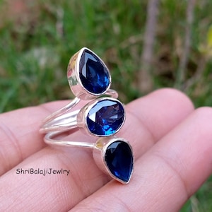 May include: A silver ring featuring three faceted dark blue stones in bezel settings. The stones are arranged in a vertical row, with a pear-shaped stone at the top, an oval stone in the center, and a smaller pear-shaped stone at the bottom.