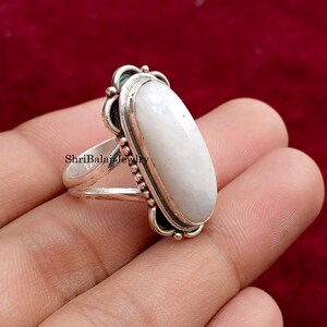 May include: A silver ring with a large, oval, white gemstone. The ring has a decorative setting with a beaded border and ornate details. The ring is held in a hand against a red background.