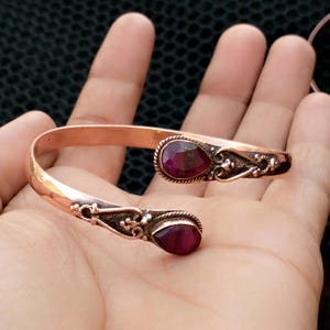 May include: A copper cuff bracelet with two oval, deep red gemstones. The bracelet has an open design with ornate, dark metal detailing around the gemstones. The bracelet is displayed on a hand.