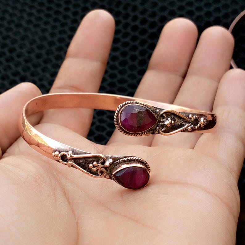 May include: A copper cuff bracelet with two oval, deep red gemstones. The bracelet has an open design with ornate, dark metal detailing around the gemstones. The bracelet is displayed on a hand.