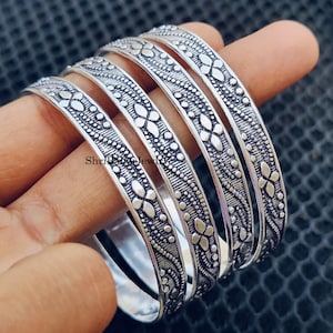 May include: A set of five silver bangles with intricate floral and dot patterns. The bangles are displayed on a hand, showcasing the detailed design and texture of the jewelry. The bangles are made of a shiny, metallic material.