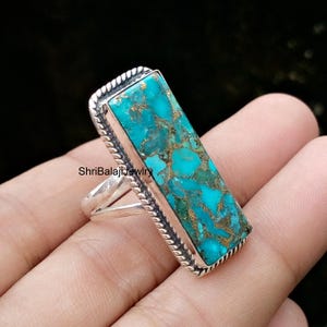Puede incluir: Un anillo rectangular con una piedra turquesa vibrante con vetas de color cobre, engastada en un marco de plata pulida con un borde de cuerda trenzada. La banda es dividida y el texto ShriBalajiJewelry es visible en el engaste de plata.