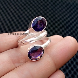 May include: A silver ring with two oval, deep purple gemstones. The ring's band wraps around, creating a unique design. The text "ShriBalajiJewlry" is visible on the ring. The gemstones have a faceted cut, enhancing their color and brilliance.