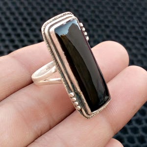 Può includere: Un anello rettangolare in argento con una grande pietra nera lucida. L'anello ha un bordo dettagliato e strutturato e piccoli accenti decorativi. La fascia è semplice ed elegante.
