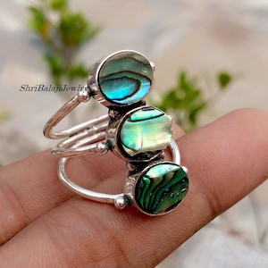 Puede incluir: Un anillo de plata con tres piedras de concha de abulón. Las piedras son ovaladas y redondas, mostrando iridiscencias azules, verdes y blancas. El anillo tiene un diseño de triple banda, con cada piedra engastada en una banda separada.