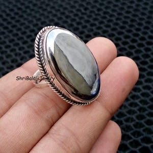 Könnte beinhalten: Ein silberner Ring mit einem ovalen, polierten, dunkelgrauen Stein. Der Ring hat einen seilartigen Rand um den Stein und ein schlichtes Band. Der Text "ShriBalajiJewlry" ist auf dem Ring sichtbar.