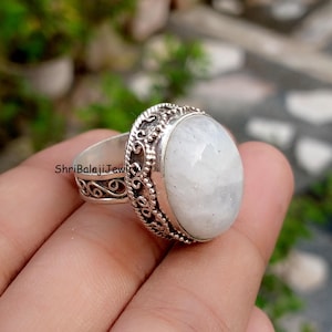 May include: A silver ring featuring a large, oval, white gemstone. The ring has an ornate, filigree design around the gemstone and on the band. The text "ShriBalajiJewl" is visible on the ring's band. The ring is set against a blurred background.