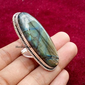 May include: A silver ring featuring a large, oval labradorite gemstone. The stone displays iridescent blue, green, and gray hues. The ring has a textured, rope-like border and the text "ShriBalajiJewelry".