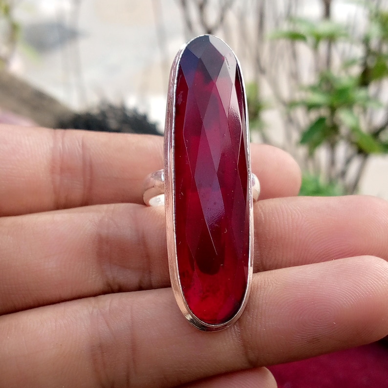 May include: A silver ring featuring a large, rectangular, faceted, deep red gemstone. The gemstone has a long, slender shape with a polished surface. The ring's band is a simple, polished silver.