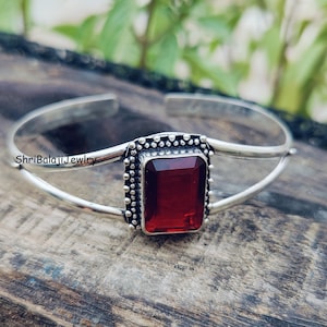 May include: A silver cuff bracelet featuring a central rectangular red gemstone. The stone is set in a frame with a beaded border. The metal band splits into two thin arms on each side. The text ShriBalajiJewelry is visible on the left side.