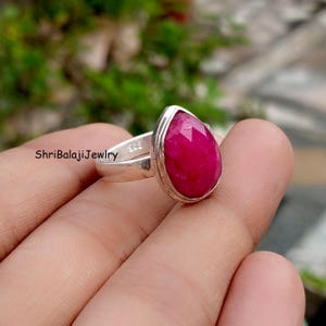 May include: A silver ring featuring a large, faceted, teardrop-shaped ruby gemstone. The ruby is a vibrant shade of red. The ring is marked with the text "ShriBalajiJewlry" and "925".