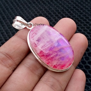 Puede incluir: Colgante ovalado con una piedra preciosa pulida en tonos rosas y morados, engastada en un marco plateado. El colgante tiene una argolla plateada para una cadena. La joya es llamativa.