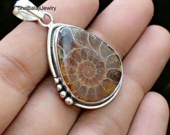 May include: A teardrop-shaped pendant with a fossilized ammonite encased in a clear, glossy material. The pendant is set in a silver-colored metal frame and has a loop at the top for a chain. The text "ShriBalajiJewlry" is visible.