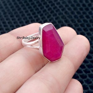 May include: A silver ring featuring a large, faceted, coffin-shaped magenta gemstone. The band is split into two thin silver segments where it meets the stone setting. The text ShriBalajiJewelry is visible on the side of the silver band.